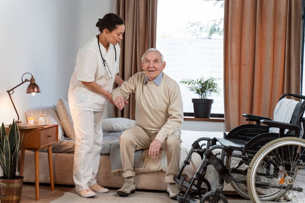 A nurse assists an elderly man to get up from a bed and to sit in a wheelchair