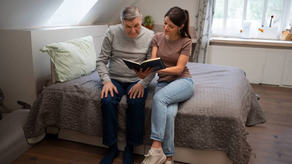 A woman and an elderly man sit on a bed, both focused on a book in their hands.