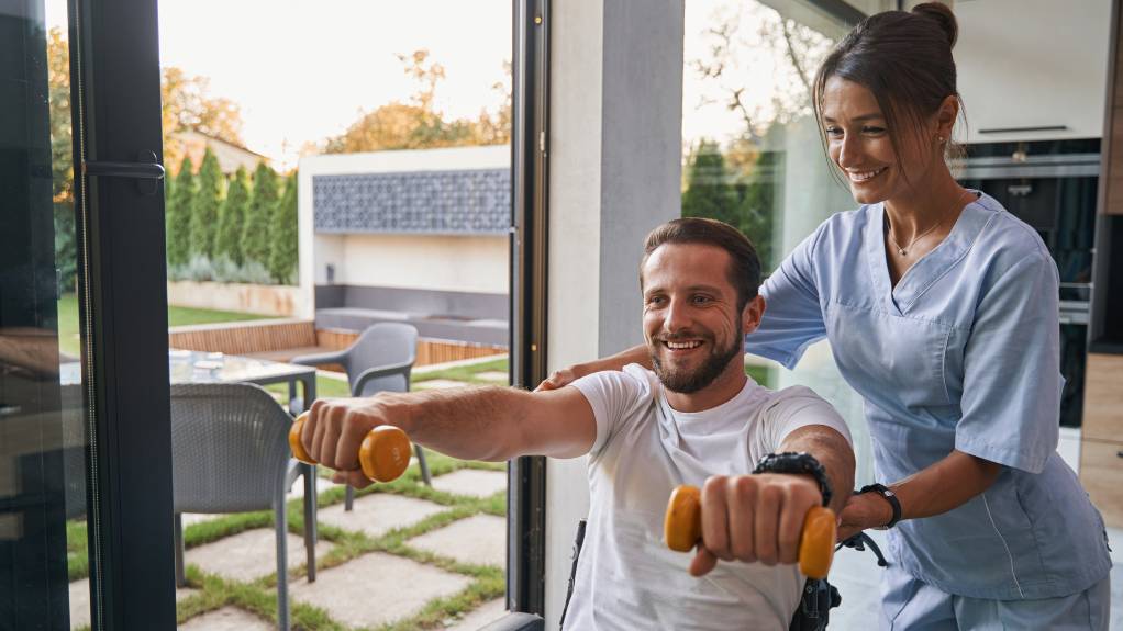 Positive male patient doing rehabilitation exercises with female physiotherapist in the room indoors